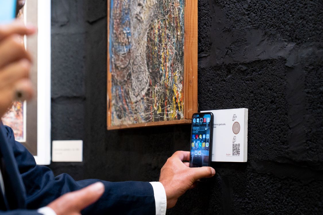 Dettaglio di una mano che avvicina lo smartphone a una targhetta con tag NFC attaccata al muro vicino a un quadro per ricevere informazioni sull’opera.