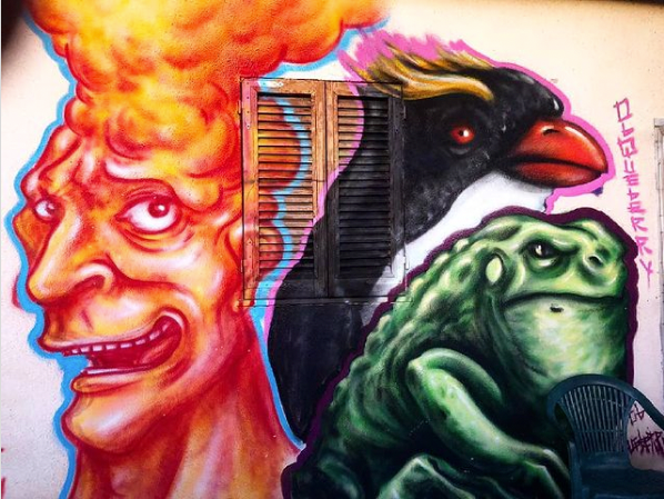 Tre opere di street art realizzate su un muro fatiscente. Da sinistra a destra: una testa umana fumante, un pinguino imperatore e un rettile simile a un’iguana.