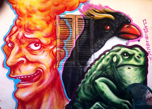 Opera di street art realizzata su un muro raffigurante una maschera con fattezze umane, caratterizzata da un lungo naso, baffi e bocca aperta in un ghigno, dalla quale sbucano i canini inferiori appuntiti. Dall’angolo in basso a destra dell’immagine spunta della vegetazione spontanea.